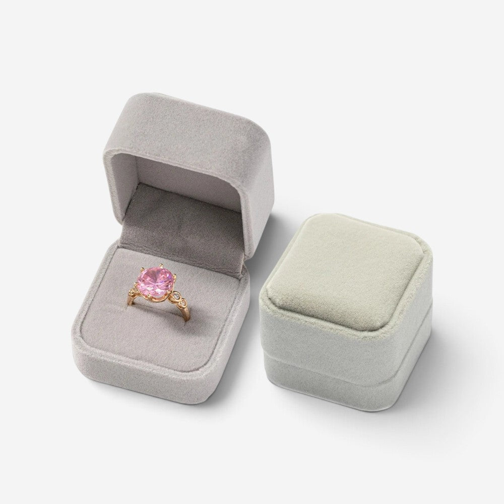 Mini boîte à bijoux SecretGleam en velours gris contenant une bague dorée avec une pierre rose